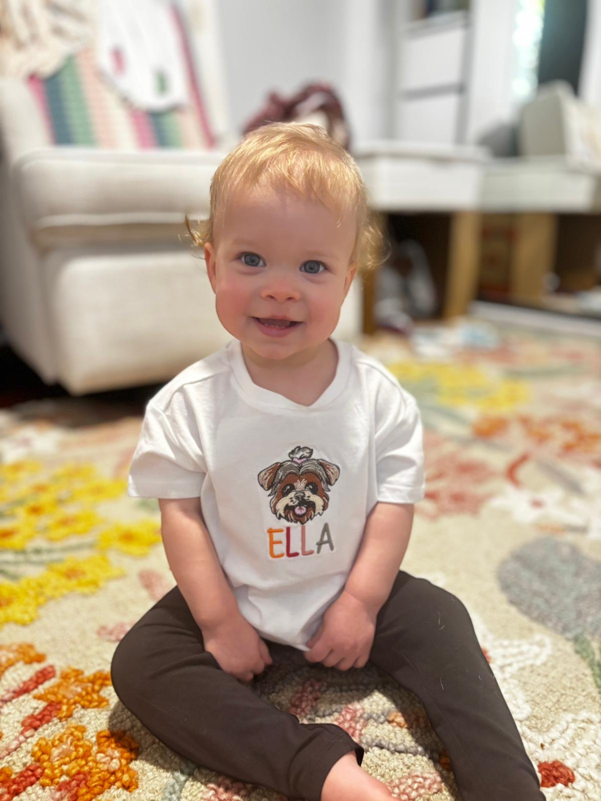 Child wearing a white shirt with a dog illustration and 'Ella' text, sitting on a colorful rug.