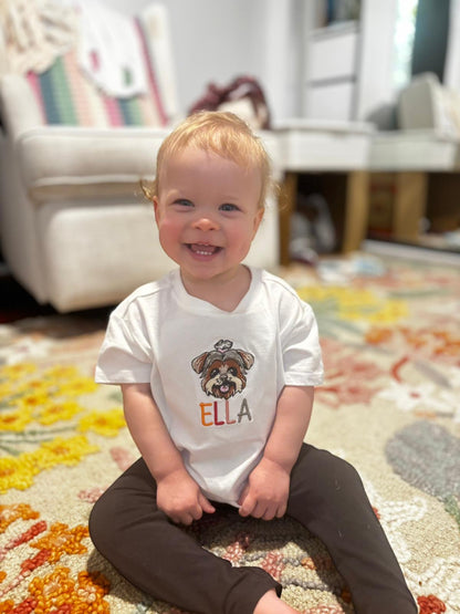 Child wearing a white t-shirt with a dog graphic and 'ELLA' text, sitting on a colorful rug.