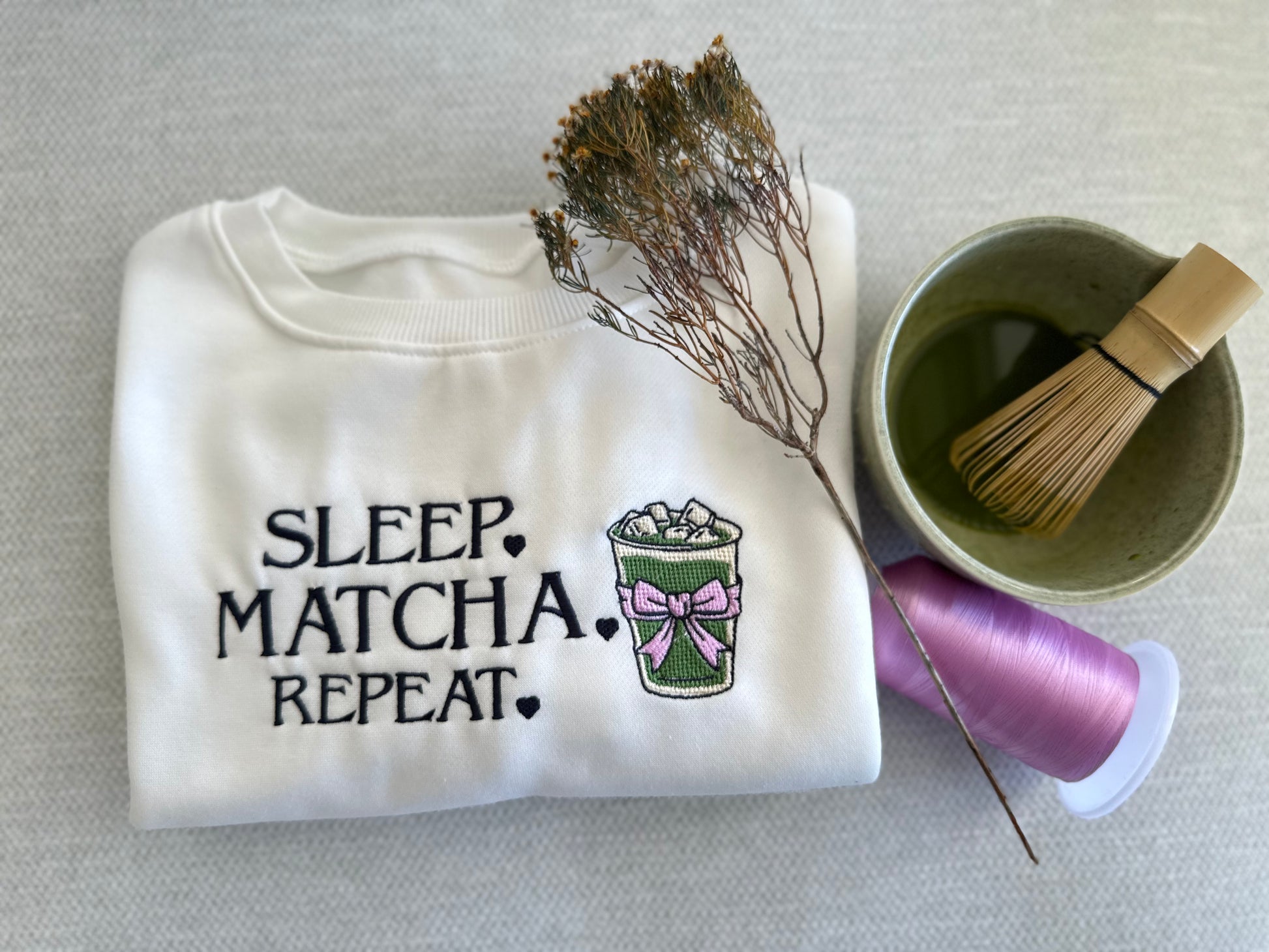 White sweatshirt with 'Sleep Matcha Repeat' text and graphic, next to a bowl with a whisk and a spool of thread on a neutral background. handcrafted embroidered apparel
cozy embroidered crewnecks
unique embroidered accessories
bookish embroidery designs
pet-themed embroidered tees
made-to-order embroidered gifts. Temu , Shein