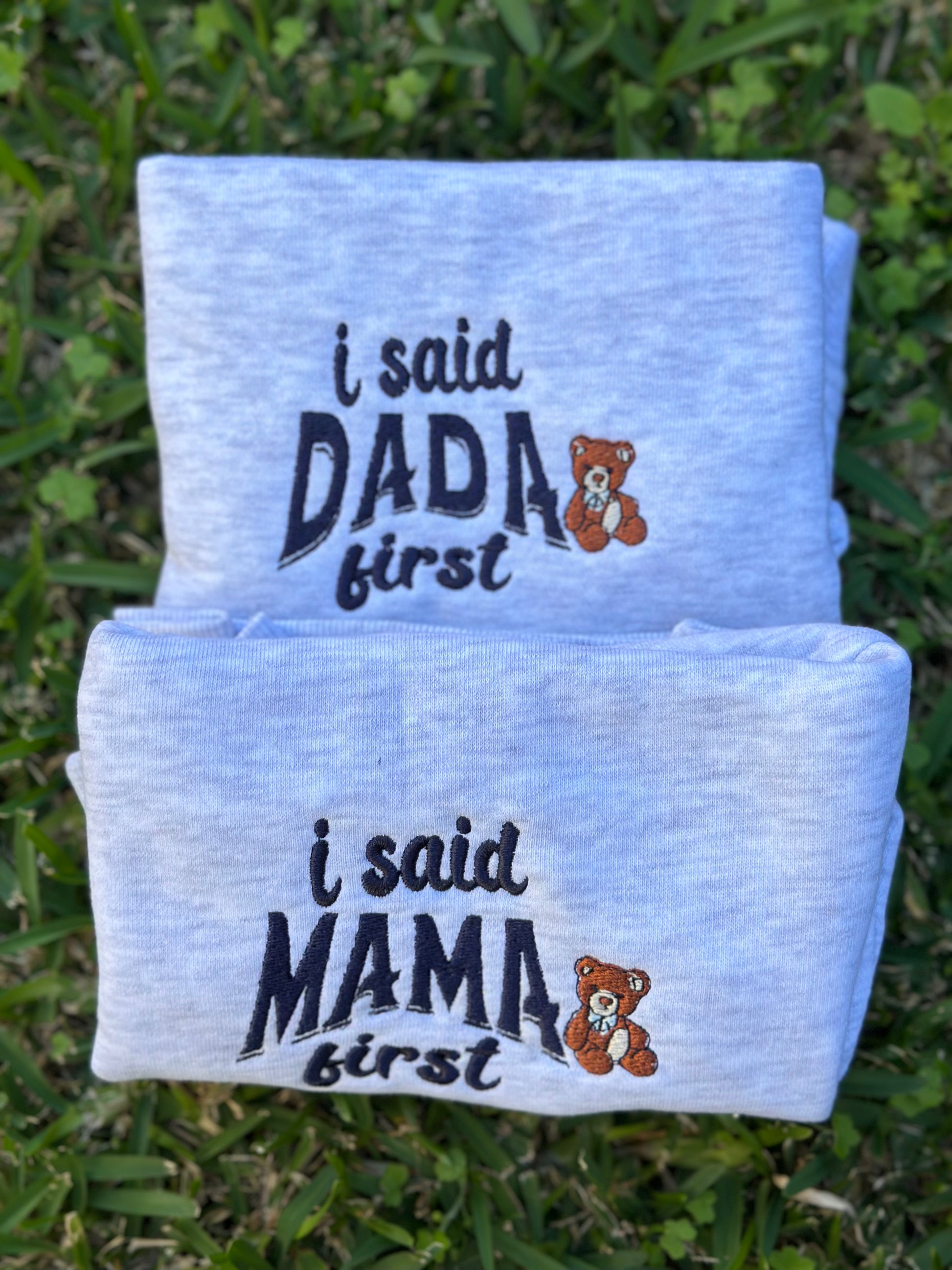 Folded baby crewneck swears with text 'i said DADA first' and 'i said MAMA first' on a grass background