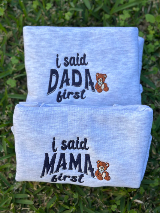 Folded baby crewneck swears with text 'i said DADA first' and 'i said MAMA first' on a grass background