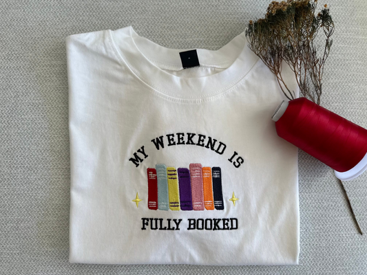 White t-shirt with colorful book design and text on a gray surface. My weekend is fully booked 